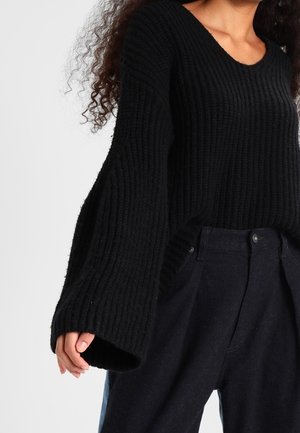Femme portant un pull en maille côtelée noire à larges manches et un pantalon noir taille haute, debout devant un fond uni.