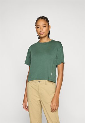 Giovane con capelli intrecciati che indossa un top corto verde a maniche corte e pantaloni beige, in piedi davanti a uno sfondo chiaro e uniforme.