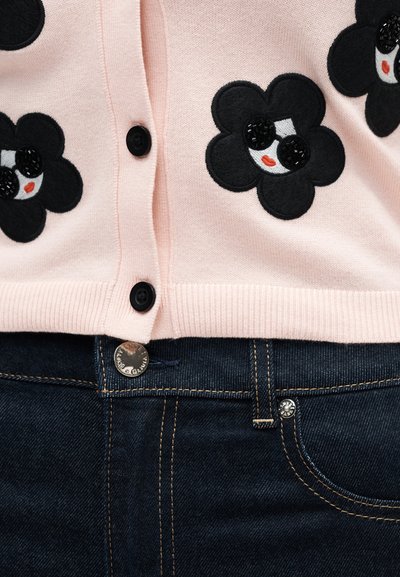 Ανοιχτό ροζ κάρντιγκαν με μαύρα μπαλώματα λουλουδιών που διαθέτουν γυαλιά ηλίου με χάντρες και κόκκινα χείλη, σε συνδυασμό με σκούρο μπλε τζιν.
