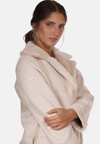 Cappotto in pelliccia sintetica beige con ampio colletto e spalle cadenti. Presenta un sottile logo dorato sul braccio sinistro. Texture morbida e vestibilità rilassata.