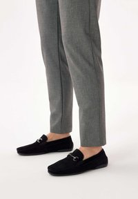 Crne antilop loafers s srebrnim detaljima, u kombinaciji s krojenim sivim hlačama. Loafers imaju ravni dizajn i minimalno šivanje.