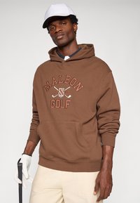 Brūns pulovers ar kapuci ar "MALBON GOLF" logo oranžā krāsā. Iekļauj kabatu kangaroo stilā un ribotas aproces. Apvienots ar krēmkrāsas biksēm un baltu cepuri.