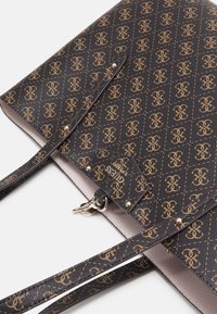 Sac cabas marron foncé avec motif logo entrelacé doré, anses fines et petite étiquette de marque Guess avec détails métalliques.