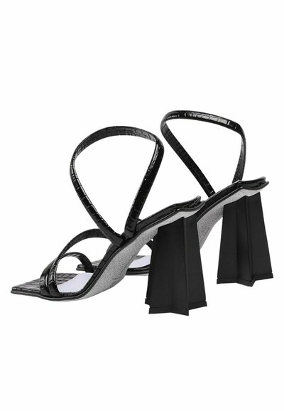 Sandali neri con tacco a stiletto in morbido materiale, caratterizzati da una texture a coccodrillo, con sottile cinturino e una forma di tacco a blocco unica. La soletta è grigia, offrendo un contrasto.