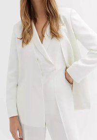 Bílý upravený blejzr s hladkým povrchem, s šálovým límcem a zakončenými rukávy, nošený přes odpovídající bílý outfit.