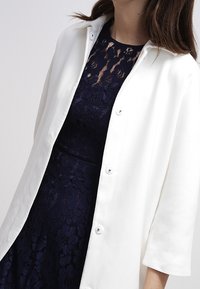 White button-up coat with a relaxed fit, featuring a collar and long sleeves, worn over a navy blue lace dress. Smooth fabric texture.