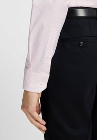 Lichtroze geruite overhemelsmouw met witte knoopmanchetten, gedragen bij donkere, gestructureerde broek met een enkele knoopdetail.