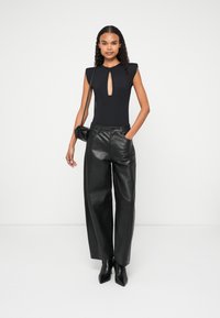 Bodysuit preto com recorte em forma de chave, combinado com calças largas de couro preto de cintura alta e sapatos de salto agudo pretos, tudo sobre um fundo branco.