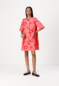 A short, flared dress in bright coral, featuring a large flower pattern. Complemented by black flats and a small pink handbag.