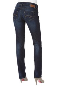 Jeans de mezclilla azul oscuro con un corte slim, que presentan bolsillos traseros curvados y un parche de cuero marrón en la cinturilla. Detalle de costura sutil en la parte delantera.
