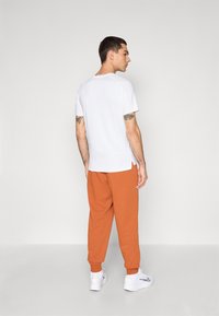 Biely tričko s krátkym rukávom v kombinácii s oranžovými teplákmi. Tepláky majú elastické manžety a celý outfit má čistý, minimalistický dizajn.