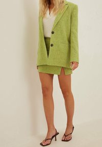 Mujer vistiendo un blazer verde lima y una falda mini a juego con una abertura lateral, top de punto blanco y sandalias de tacón marrón frente a un fondo liso.