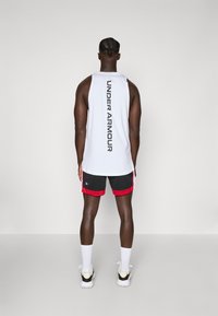 Débardeur de sport sans manches blanc avec "UNDER ARMOUR" imprimé verticalement au dos, associé à un short en mesh noir et rouge.