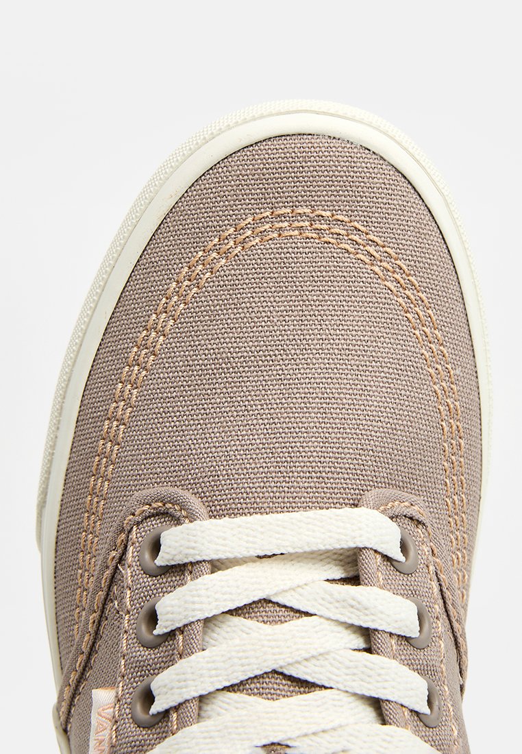 Zapatillas de lona marrones con puntera redondeada, cordones blancos y costuras de un beige claro. Presentan una parte superior texturizada y suela de goma para mayor agarre.