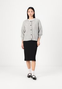 Grå strikket cardigan med rund hals, seks mørke knapper foran og ribbestrikkede mansjetter, kombinert med et svart knelangt skjørt.