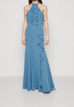 Woman wearing a floor-length blue halter dress with ruffled details on the neckline and side, standing against a plain light background.