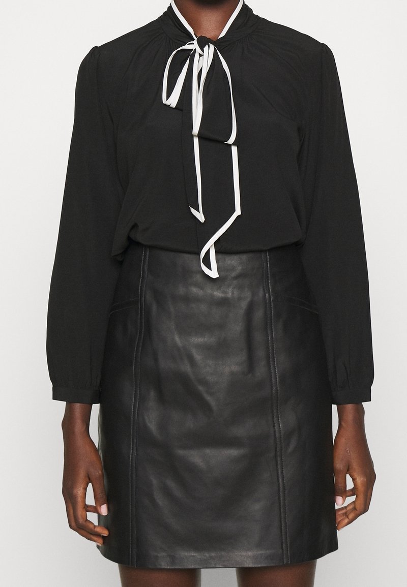 Schwarze Bluse mit langen Ärmeln, gerafftem Kragen und weißen Bindebändern, kombiniert mit einem schwarzen Leder-Minirock mit seitlichen Taschen und vertikalen Nähten.