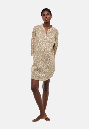 Femme debout pieds nus portant une robe beige à fleurs arrivant aux genoux, avec des manches longues et un col en V fendu, sur fond blanc.