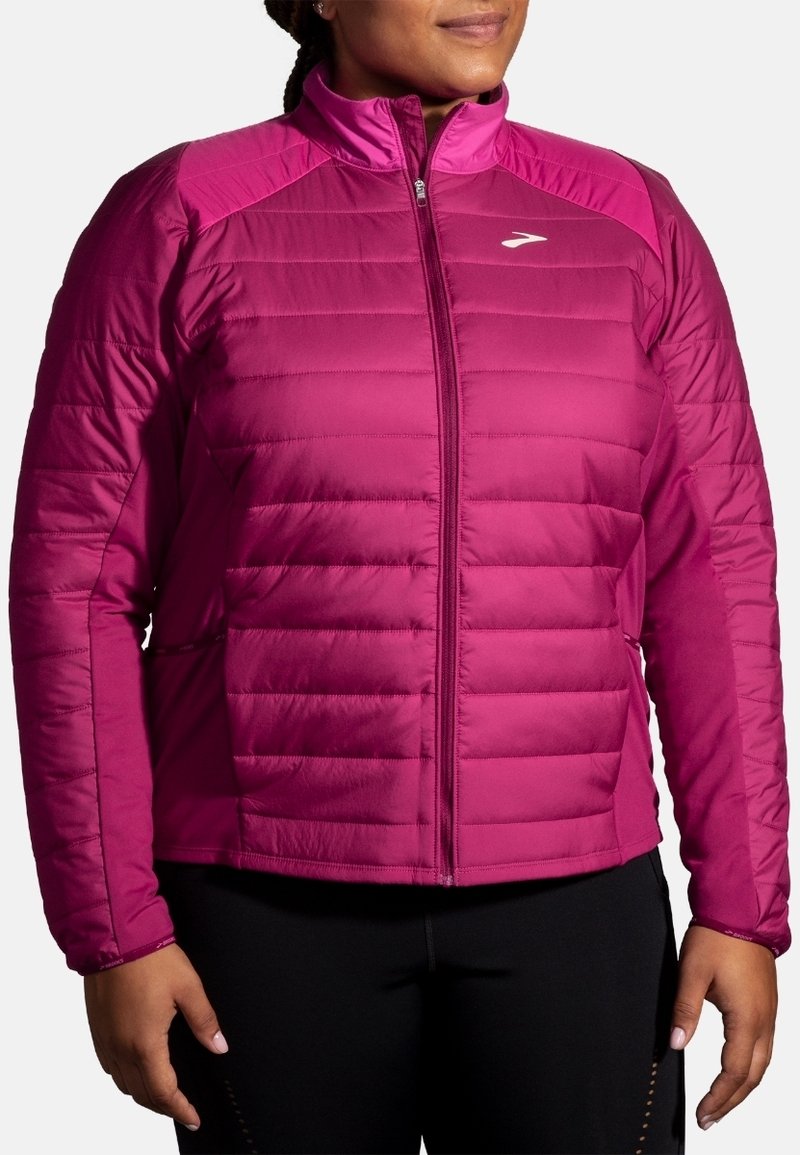 Femme portant une veste matelassée magenta à fermeture éclair, à manches longues, et un pantalon noir, debout les mains détendues le long du corps.