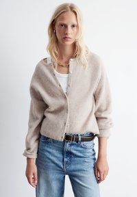 Beige gestrickte Strickjacke mit einer vorderen Knopfleiste, geripptem Muster und umgeschlagenen Ärmeln, kombiniert mit hellblauen, hoch taillierten Jeans und einem braunen Gürtel.