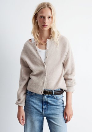 Beige knitted cardigan with a front button closure, ribbed texture, and rolled sleeves, paired with light blue high-waisted jeans and a brown belt.