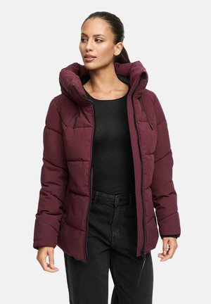 Vrouw in een bordeauxrood gewatteerd pufferjack met een grote capuchon, over een zwart top en zwarte broek, kijkend opzij.