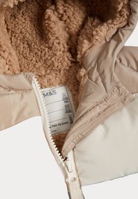 Beige quiltad jacka med ett mjukt beige fleecefoder. Synlig dragkedja och skötselråd inuti. Slät ytstruktur.