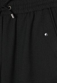 Zwarte joggerbroeken van zacht materiaal hebben een elastische tailleband, een trekkoord en drukknoopdetails op de zakken.