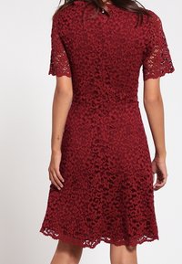 Maroon lace-mekko, jossa on lyhyet hihat, kukkakuosi, istuva yläosa, levenevä hame ja aallotettu helma. Takaa nähtynä yksityiskohdat ja kuosi.