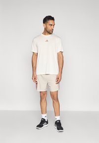 Μπεζ αθλητικό t-shirt με μικρό καφέ λογότυπο, σε συνδυασμό με ανοιχτόχρωμο σορτς με κόκκινες ρίγες στο πλάι, μαύρα αθλητικά παπούτσια και λευκές κάλτσες.