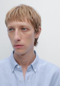 Light blue button-up shirt with a collar, featuring a smooth texture and small white buttons. Model has short blonde hair and a neutral expression.