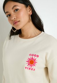 Jeune femme aux longs cheveux noirs portant des boucles d'oreilles en or et un sweat-shirt crème avec un motif de soleil rose et orange ainsi que le texte "GOOD VIBES".