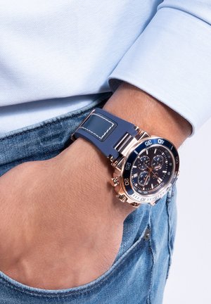 Blauwe rubberen horlogeband met een roségouden kast, zwarte wijzerplaat en witte accenten; beschikt over chronograaf sub-dials en luminescente wijzers.