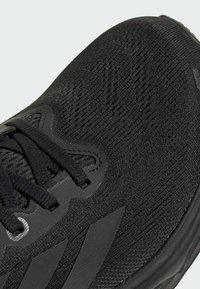 Scarpa sportiva nera con tomaia a maglia, superficie texture e suola in gomma. Caratteristiche includono lacci e dettagli del logo.