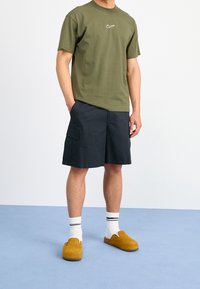 Green cotton t-shirt with white logo, black cargo shorts, white socks with black stripes, and brown suede slip-on shoes.
