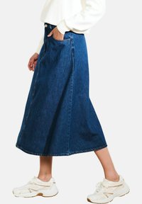 Donkerblauwe denim midi-rok met een uitlopende snit, voorzakken en een rafelige zoom. Gecombineerd met witte sneakers voor een casual uitstraling.