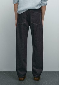 En person står vendt væk fra beskueren, iført mørke jeans og en lys blå skjorte med knapper mod en neutral baggrund.