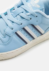 Blå lädersneaker med tre vita och marinblå ränder, texturerade skosnören och en vit gummisula med sömdekor.