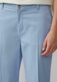 Pantaloni azzurri chiari realizzati in tessuto morbido, con chiusura a bottone, tasche frontali e gambe rette. Mano appoggiata sul tessuto.