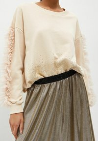Mujer vestida con un suéter beige de mangas acampanadas y pequeños detalles en forma de perlas, combinado con una falda metálica plisada con cintura negra.