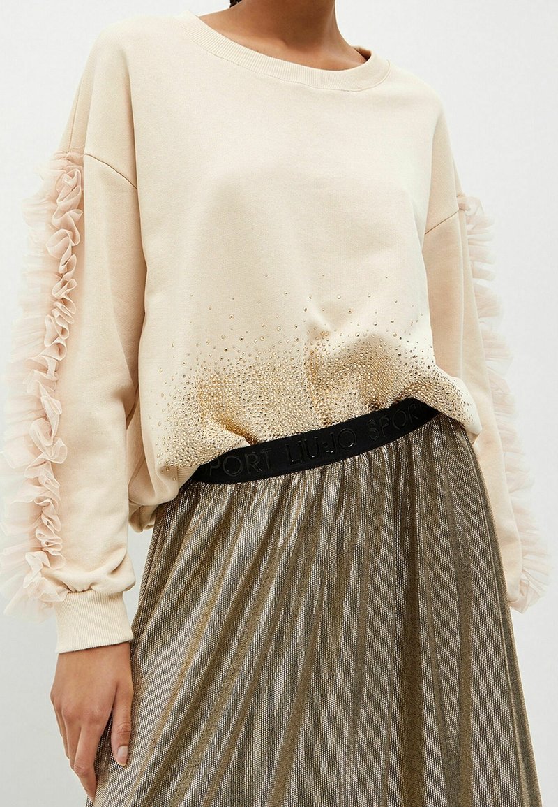 Mujer vestida con un suéter beige de mangas acampanadas y pequeños detalles en forma de perlas, combinado con una falda metálica plisada con cintura negra.
