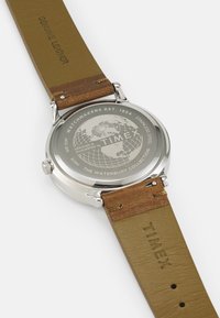 Caja de reloj de acero inoxidable plateado con una correa de cuero genuino marrón. La parte trasera presenta un diseño de globo y la marca Timex.