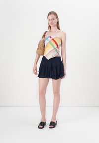 Haut multicolore à une épaule avec des rayures diagonales, jupe nouée marine, sac en osier marron, et chaussures à glisser noires. Design simple et moderne.