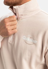 Light pink quarter-zip pullover made of soft cotton blend. Features a white logo on the chest and ribbed collar, with a smooth texture.