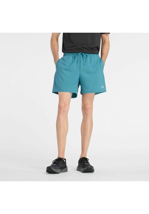 Mann trägt türkise Sportshorts und schwarze Sneakers, steht mit den Händen in den Taschen vor einem schlichten weißen Hintergrund.