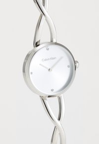 Montre en métal argenté avec un cadran rond blanc, trois aiguilles minimalistes et quatre marqueurs d'heure circulaires ; présente un design de bracelet torsadé.