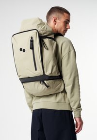 Beige Rucksack mit schwarzen Akzenten, rechteckiger Form, verstellbaren Trägern und einem Klappverschluss oben. Verfügt über eine Reißverschlusstasche auf der Vorderseite.