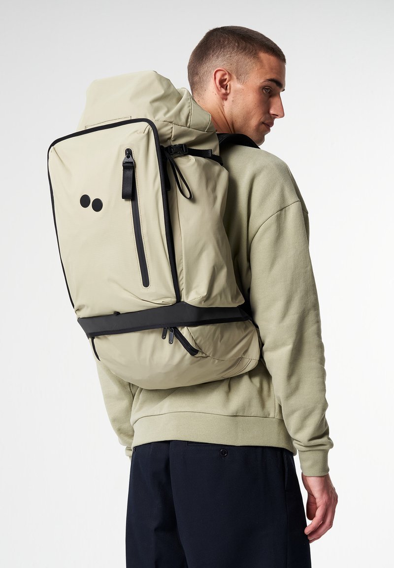 Beige Rucksack mit schwarzen Akzenten, rechteckiger Form, verstellbaren Trägern und einem Klappverschluss oben. Verfügt über eine Reißverschlusstasche auf der Vorderseite.