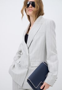 Lichtgrijze pinstripe blazer met dubbele knopen, een marineblauwe clutch met gestikte accenten, gedragen over een donkere top.