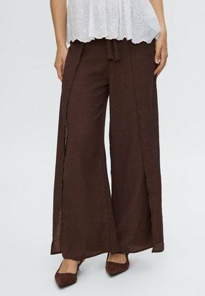Mujer vistiendo pantalones anchos de color marrón oscuro con aberturas laterales, combinados con zapatos marrón oscuro de punta afilada y una blusa blanca con ojalillos.